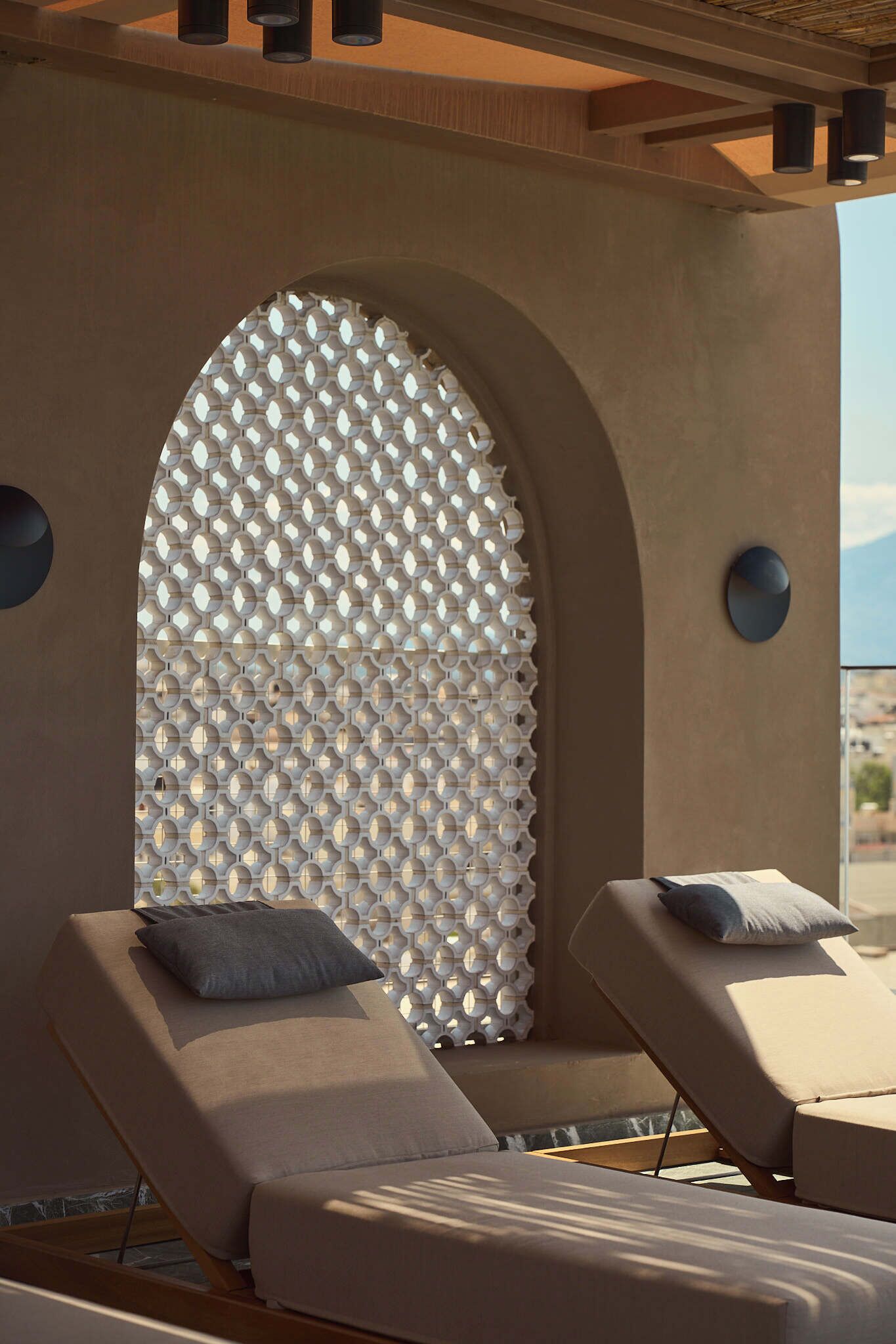A sunlit outdoor lounge area with two reclining chairs, a textured wall, and a large arched window.