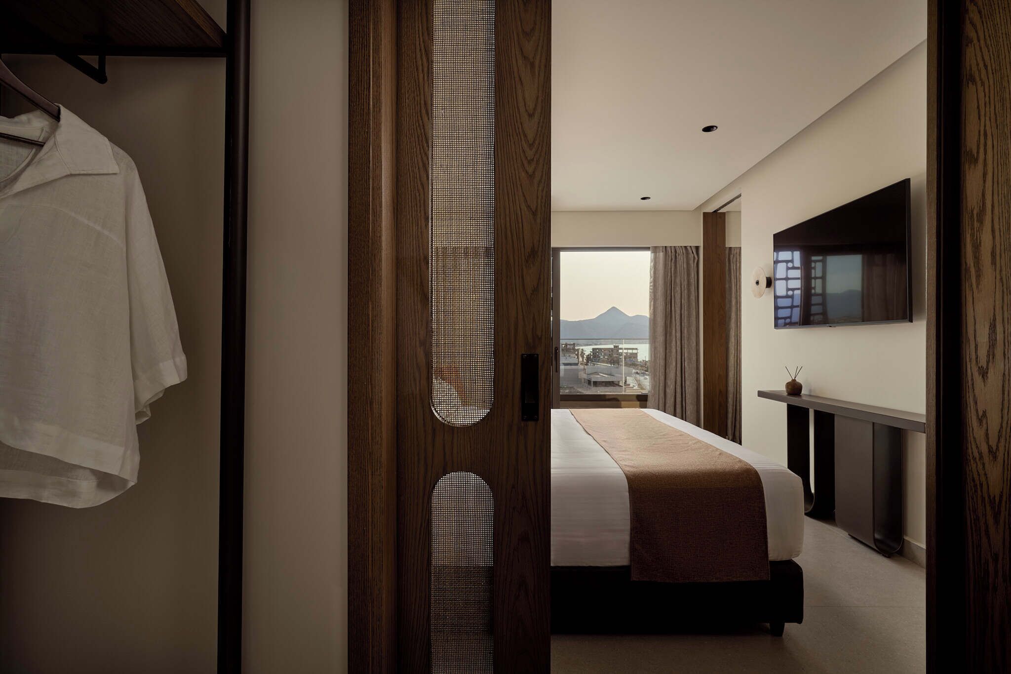 A hotel room with a bed, a TV, a window with a mountain view, and a wooden door.