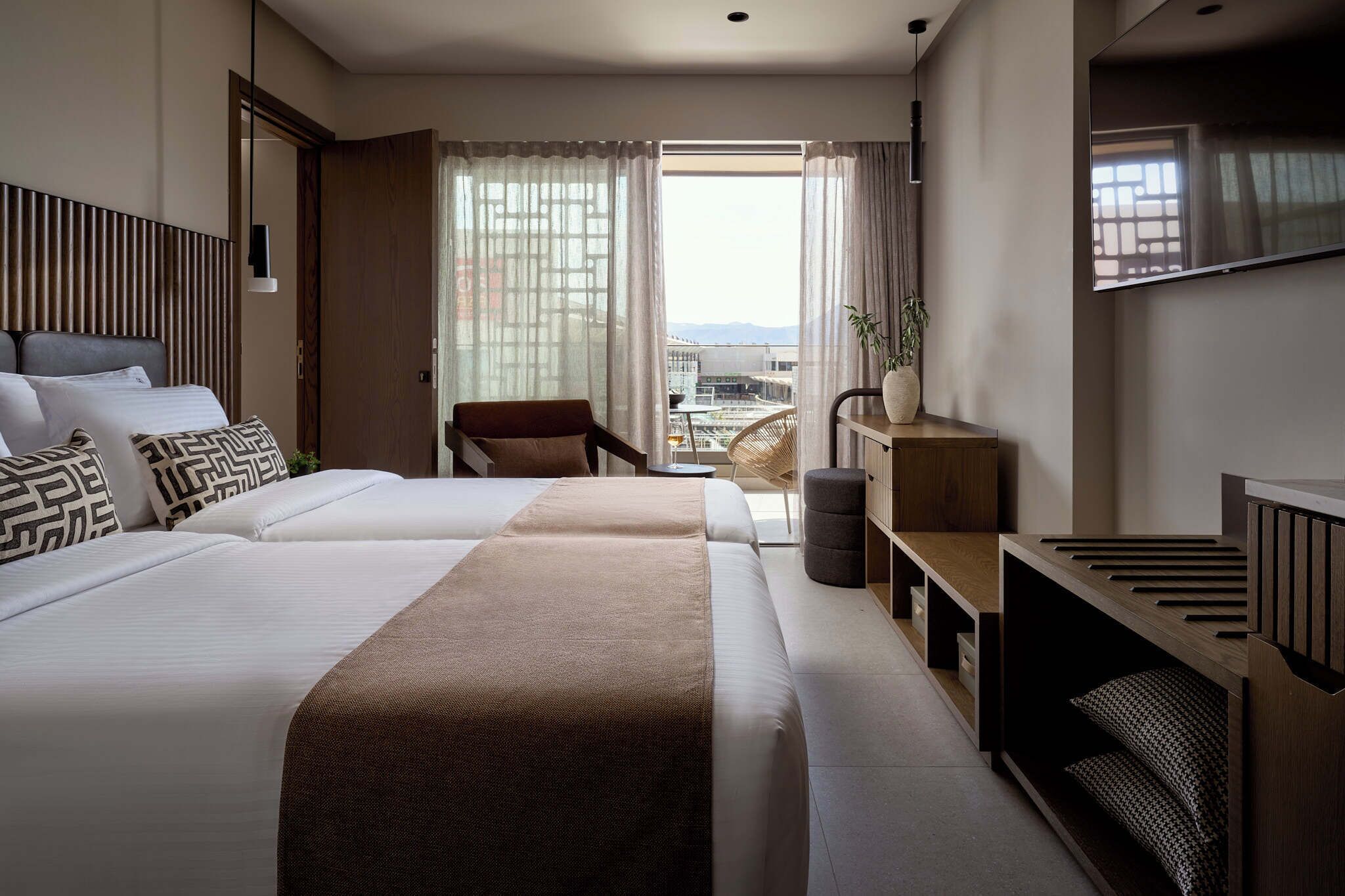 A hotel room with a large bed, a chair, a TV, and a balcony with a view of the city.