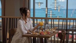 A person seated at a table with a meal and a drink, overlooking a beach with the ocean and a red car in the background.