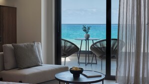 A modern living room with a sofa, two wicker chairs, a small table with a bowl and book, and a view of the ocean through a sliding glass door.