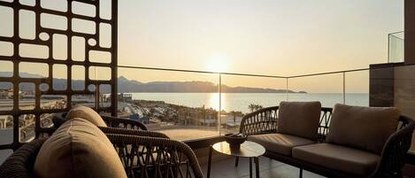 Een balkon met rieten meubels, een glazen scherm en uitzicht op zee en bergen bij zonsondergang.
