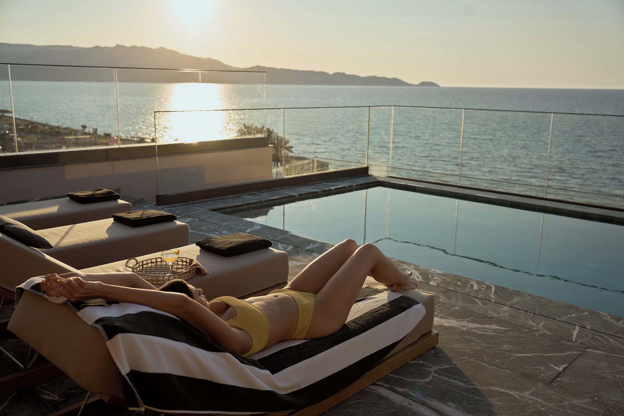 Una persona descansando en una tumbona junto a una piscina con vista al mar y a las montañas.