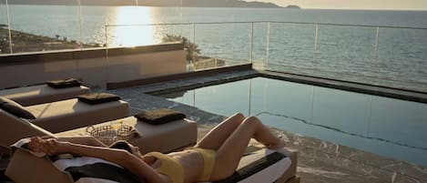 A person relaxing on a sun lounger by a pool with a view of the sea and mountains.
