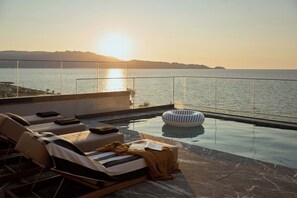 Une piscine à débordement avec des chaises longues et une vue sur la mer au coucher du soleil.