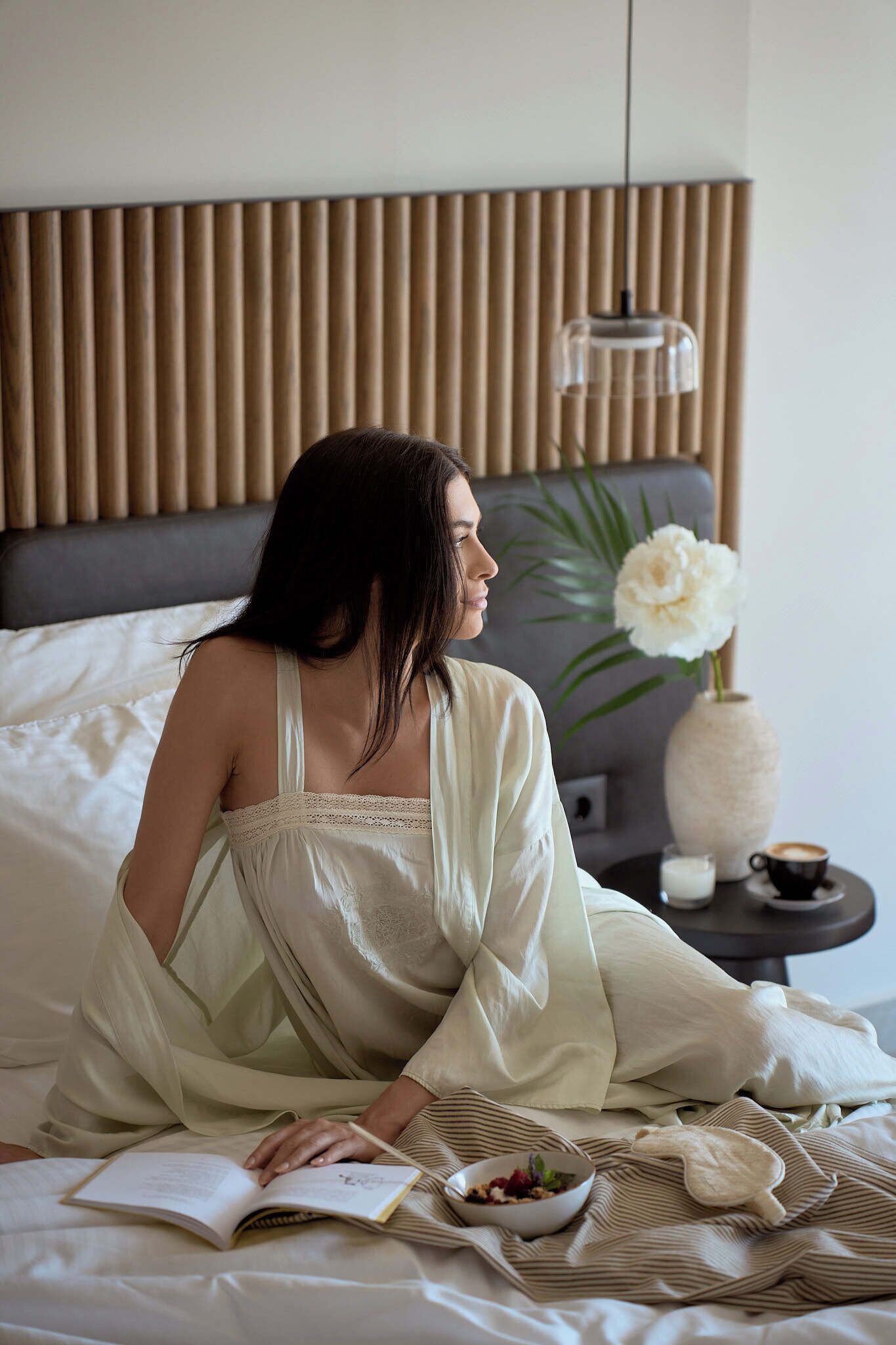 A person sitting on a bed with a book, a bowl of fruit, and a vase with a flower.