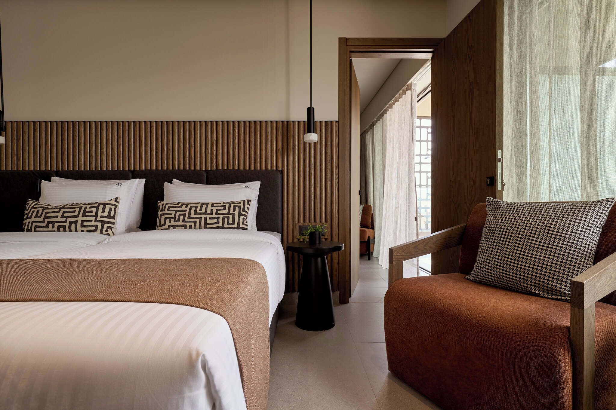 Ein modernes Hotelzimmer mit einem großen Bett, einem kleinen Beistelltisch, einem Holzstuhl und Blick auf den Korridor.