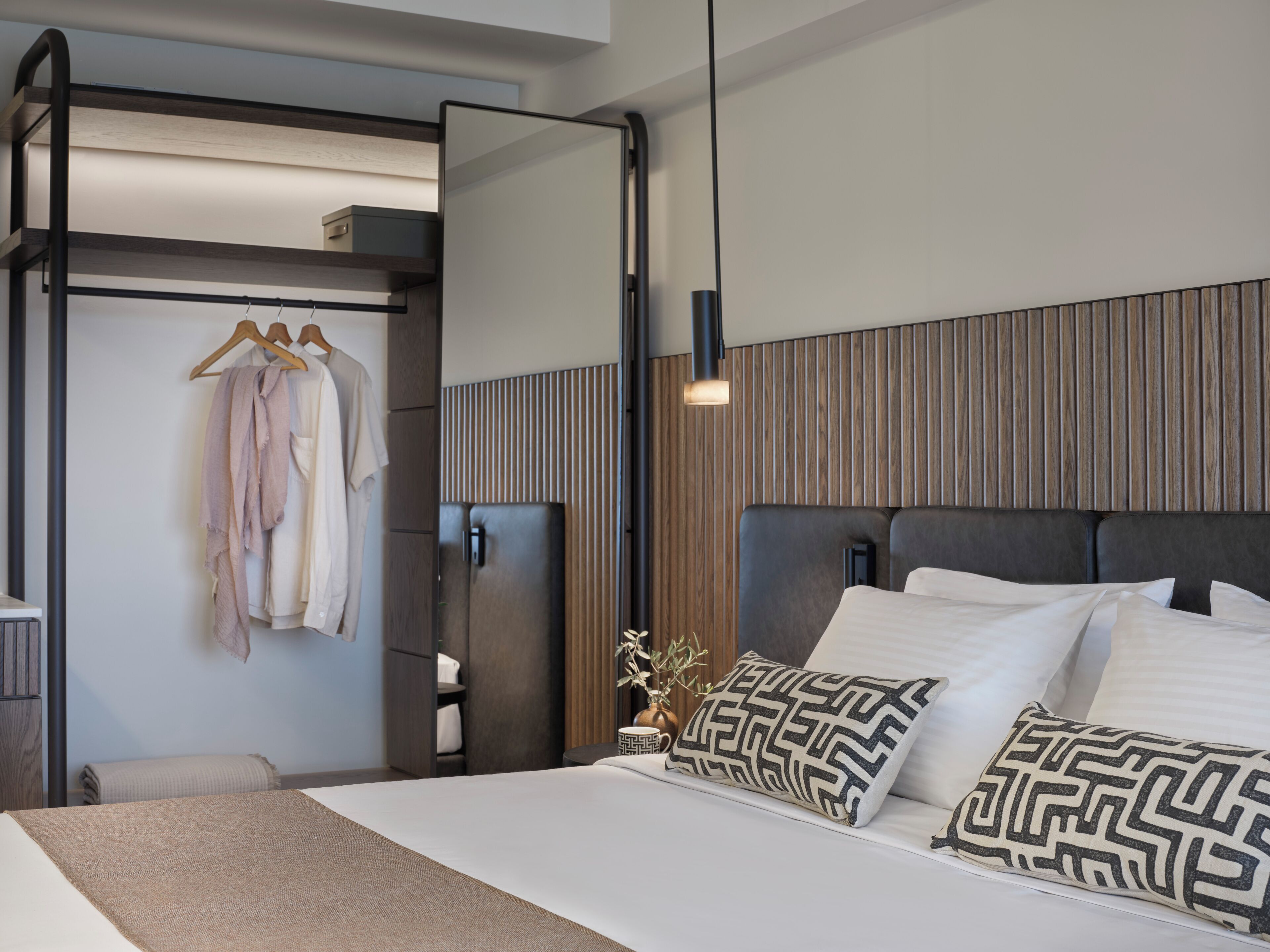 A modern hotel room with a large bed, a wardrobe with hanging clothes, a bedside table, and a mirror.