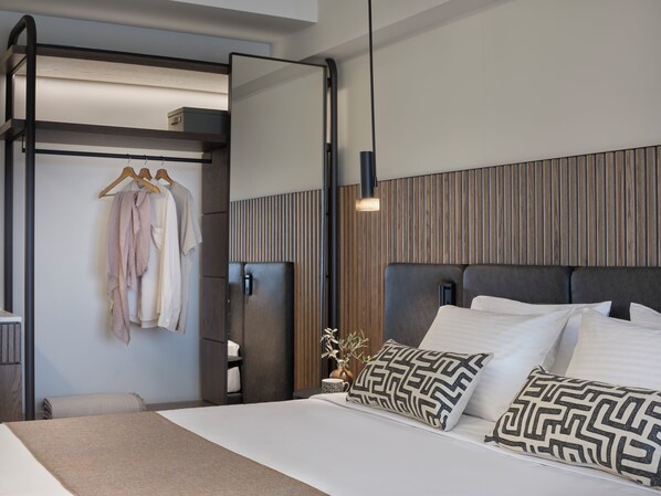 A modern hotel room with a large bed, a wardrobe with hanging clothes, a bedside table, and a mirror.