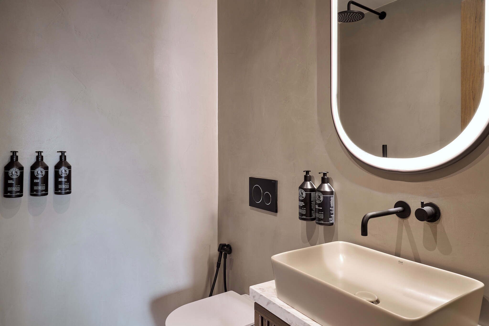 Een badkamer met een grote, ovale spiegel, een beige wastafel en drie zwarte flessen aan de muur.