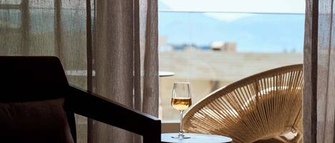 A room with a view of the sea, a wicker chair, and a glass of wine on a small table.