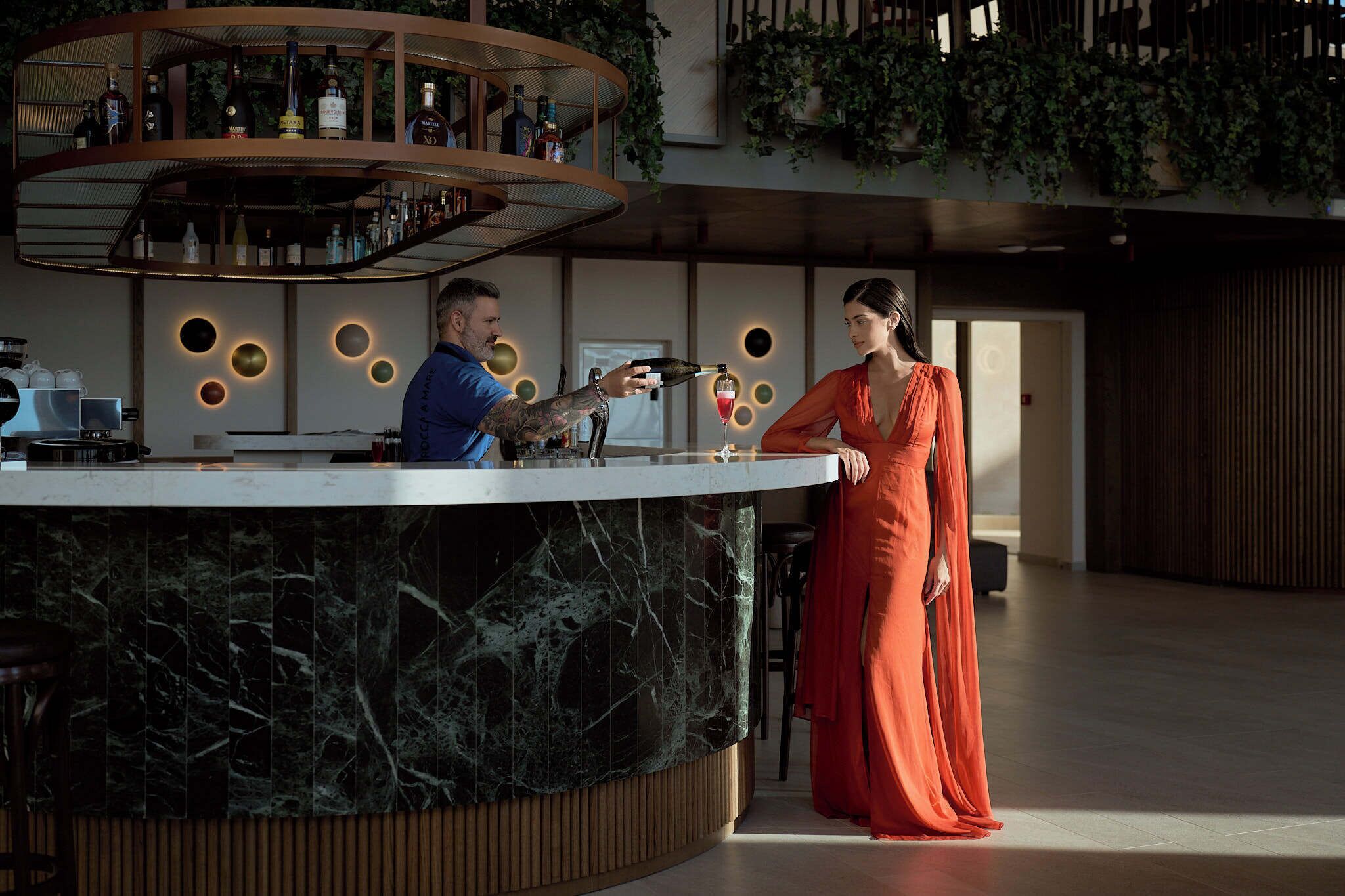 Een moderne bar met een rond bord voor flessen, een marmeren aanrecht en een vrouw in een rode jurk die er staat.