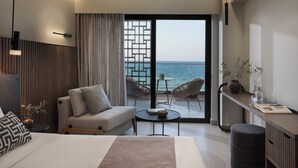 A modern hotel room with a bed, a sofa, a small table, and a balcony with a view of the sea.