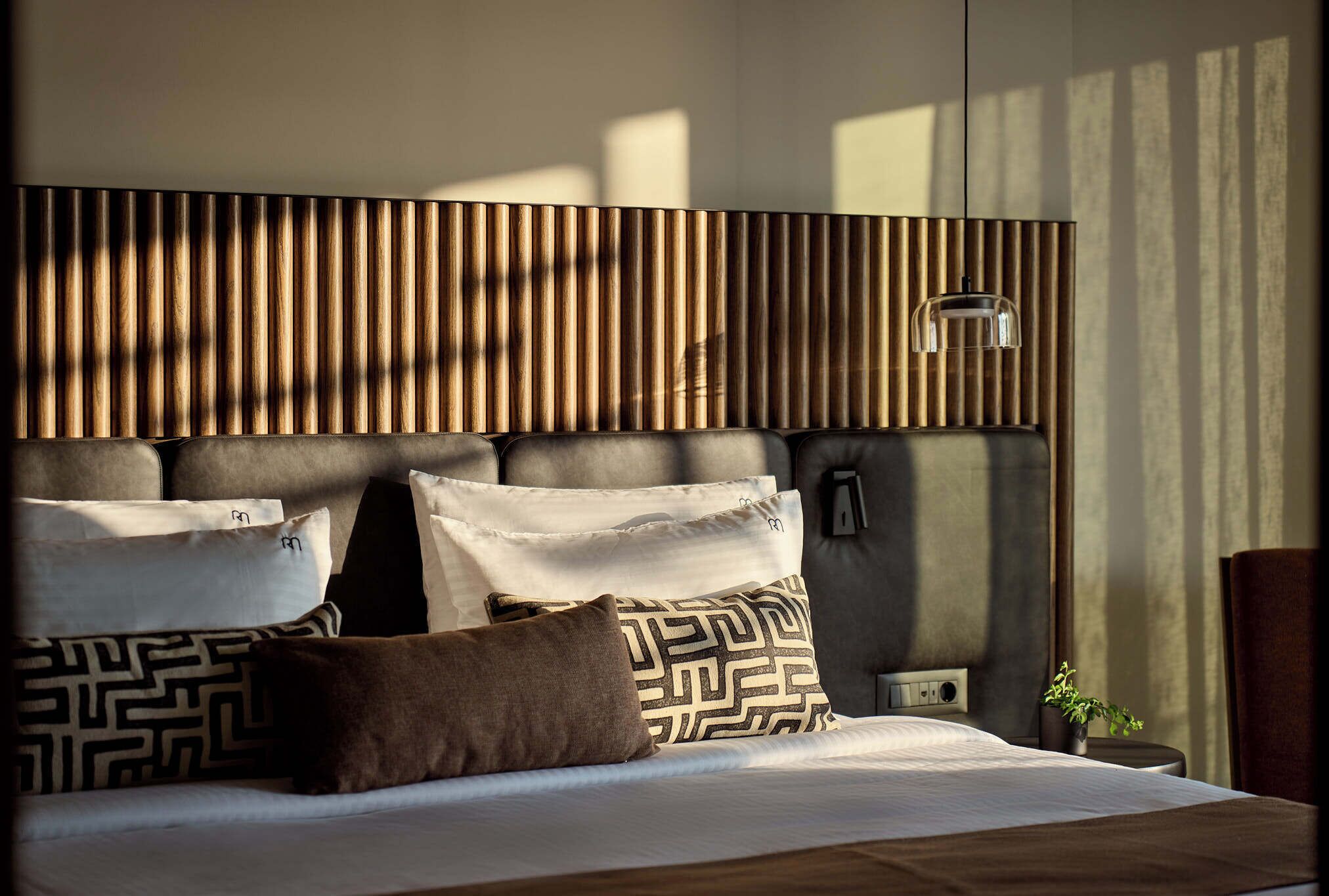 Una habitación de hotel moderna con un cabecero de madera, una cama con almohadillas estampadas, una mesita de noche con lámpara y una planta pequeña.