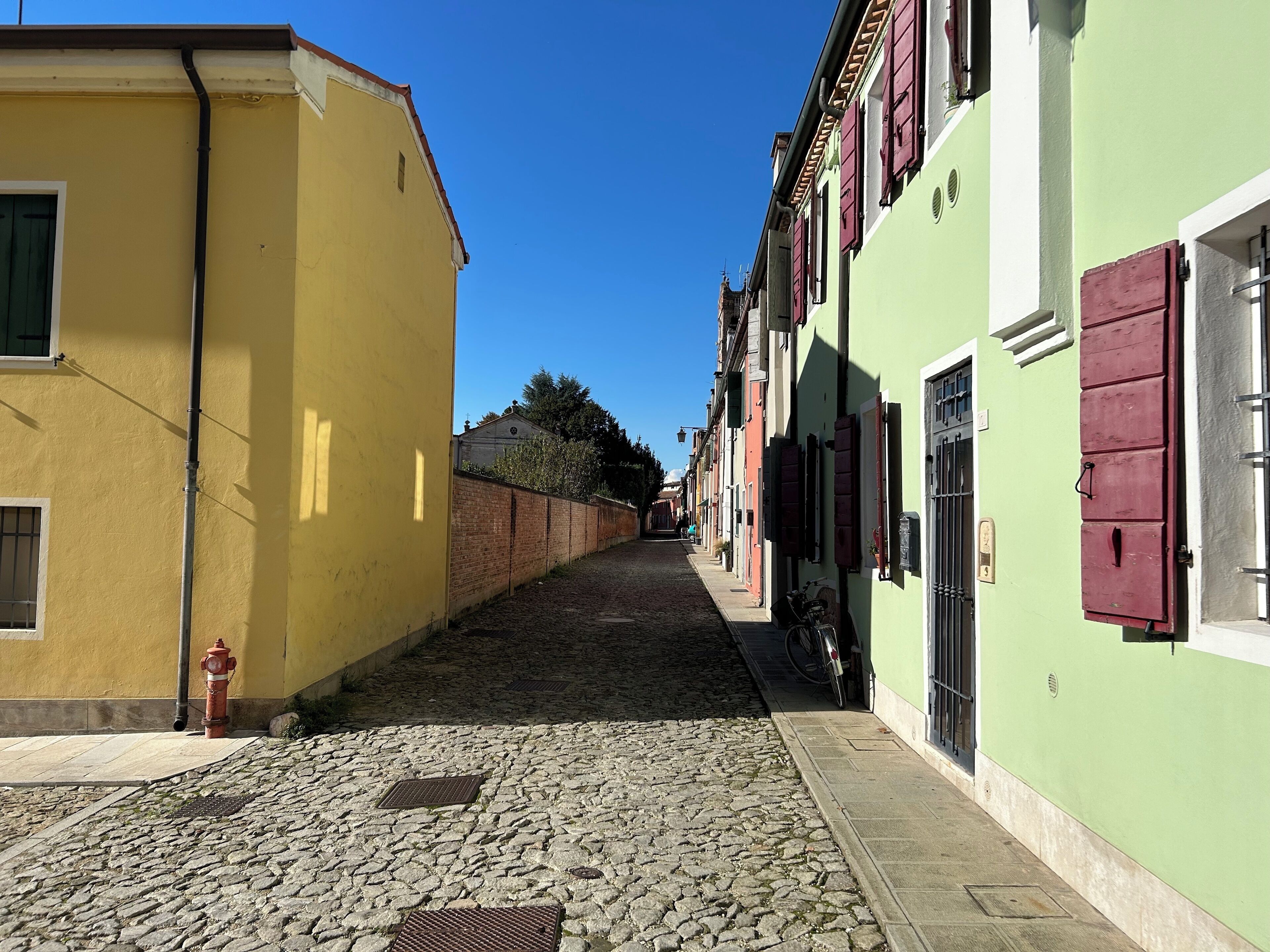 A narrow cobblestone street flanked by buildings with shutters, a fire hydrant, and a clear blue sky.
