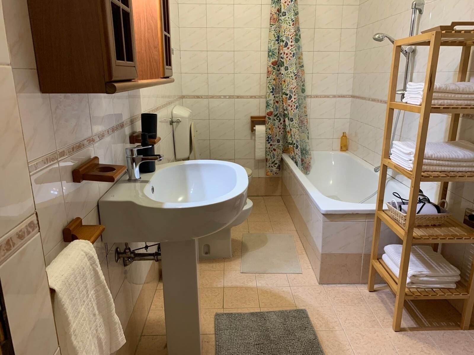 A bathroom with a sink, toilet, bathtub, wooden shelf with towels, and a shower curtain.