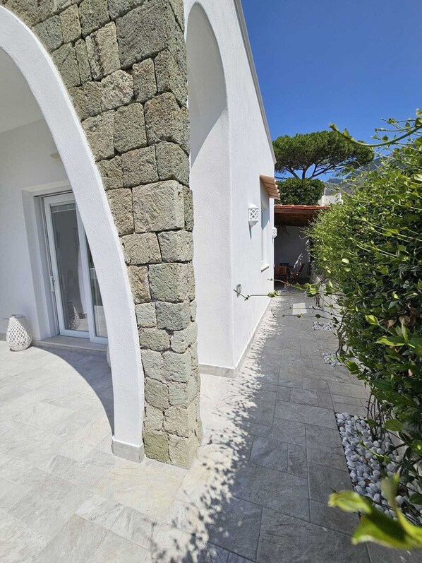 A building with stone walls and white arches, a tiled pathway, and a garden with shrubs and a tree.