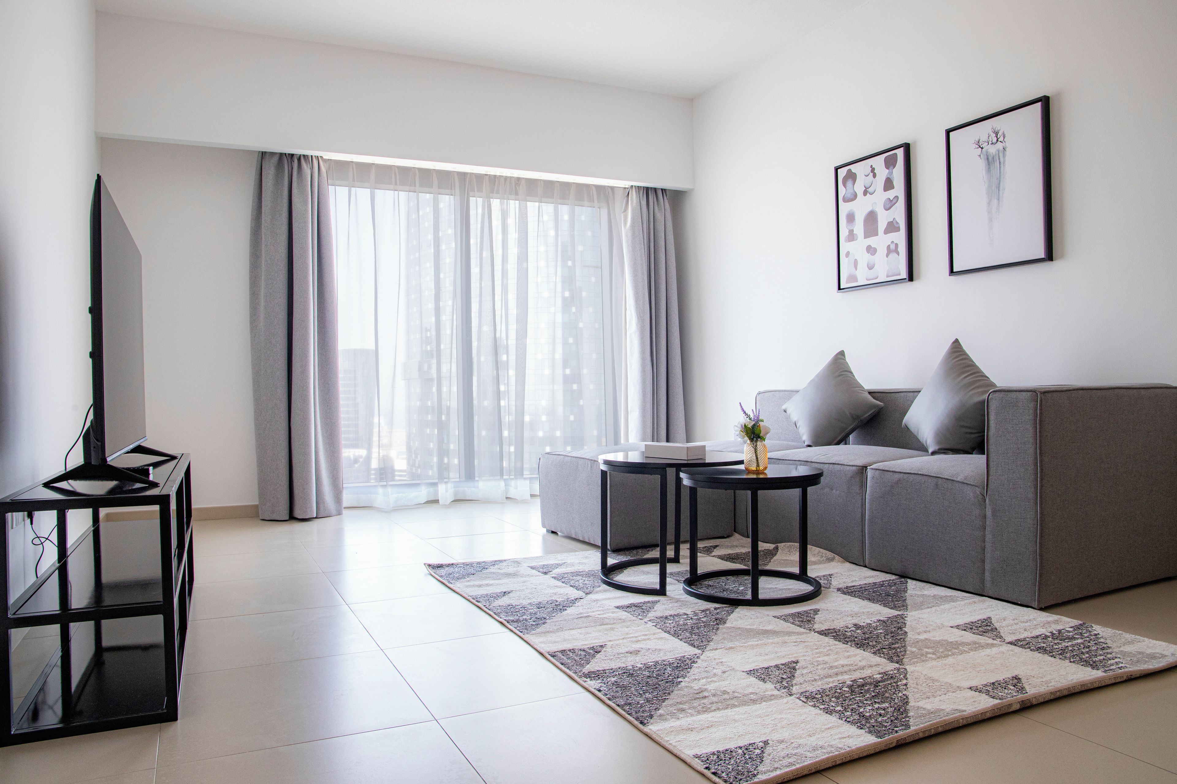 A modern living room with a grey sofa, two black side tables, a flat-screen TV, and a large window with sheer curtains.