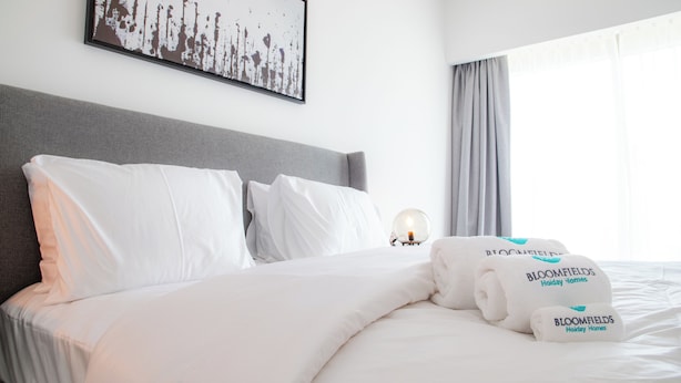 Una cama bien hecha con sábanas y almohadas blancas, un cabecero gris, una obra de arte enmarcada en la pared y una ventana con cortinas.