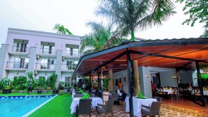 An outdoor dining area with tables and chairs, a pool, and a building with balconies. - The Mansion Hotel (Jinja)