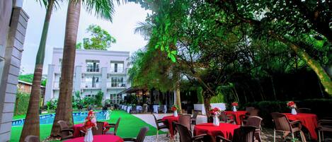 An outdoor dining area with tables and chairs, a pool in the background, and a building with balconies.