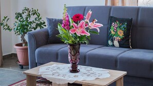 En stue med en grå sofa, et træbord med hvide blonde og en vase med blomster, samt en potteplante.