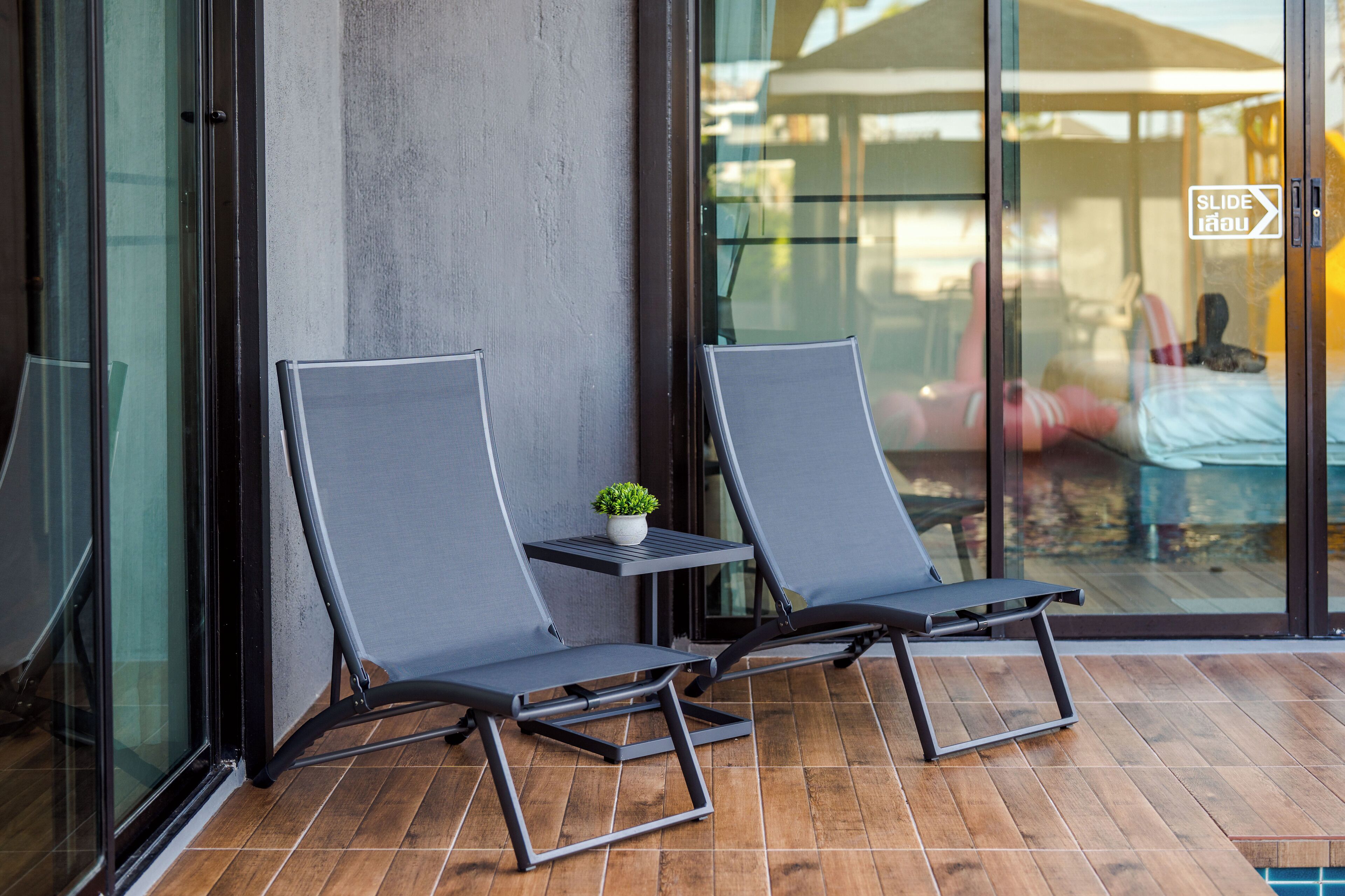 Two lounge chairs with a small table and a potted plant between them, positioned on a wooden deck with a glass wall in the background.