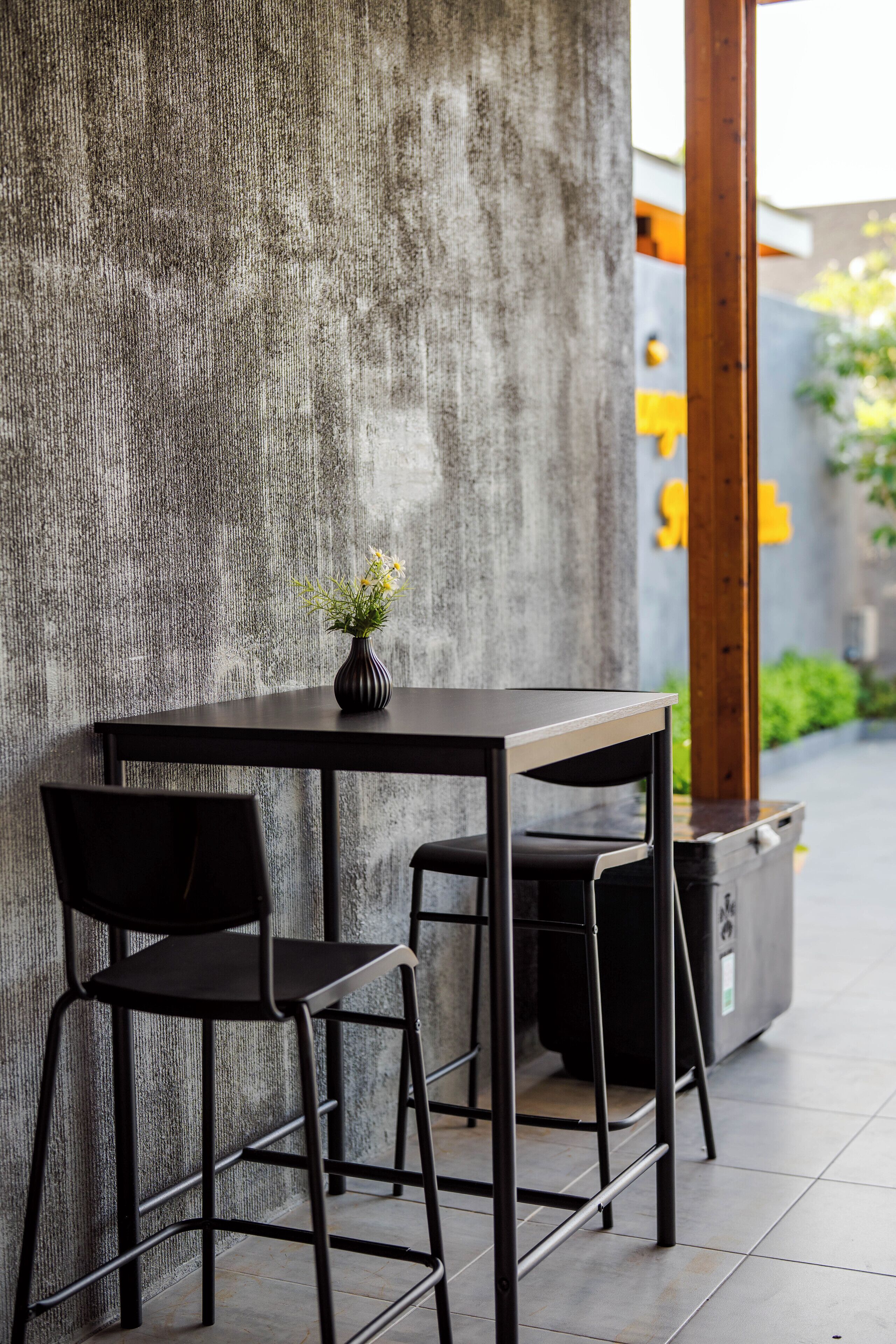 A modern outdoor seating area with a concrete wall, a black table, two black chairs, and a potted plant.