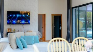 Ein modernes Wohnzimmer mit einem an der Wand montierten Flachbildfernseher, einem Sofa mit blauen Kissen, einem Ventilator und einem Essbereich mit Tisch und Stühlen.