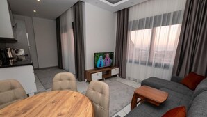 A modern living room with a grey sofa, a wooden coffee table, and a TV on a wooden stand. There is a dining area with a round wooden table and chairs.