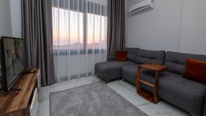 A modern living room with a grey sectional sofa, a wooden side table, and a large window with sheer curtains.