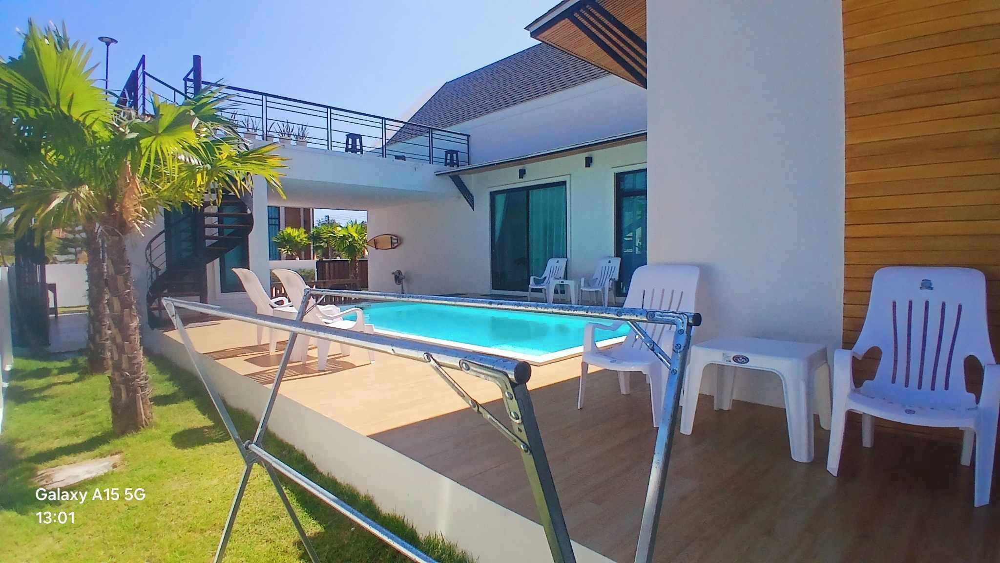 Un área de piscina exterior moderna con barandilla de vidrio, sillas reclinables blancas y cubierta de madera.