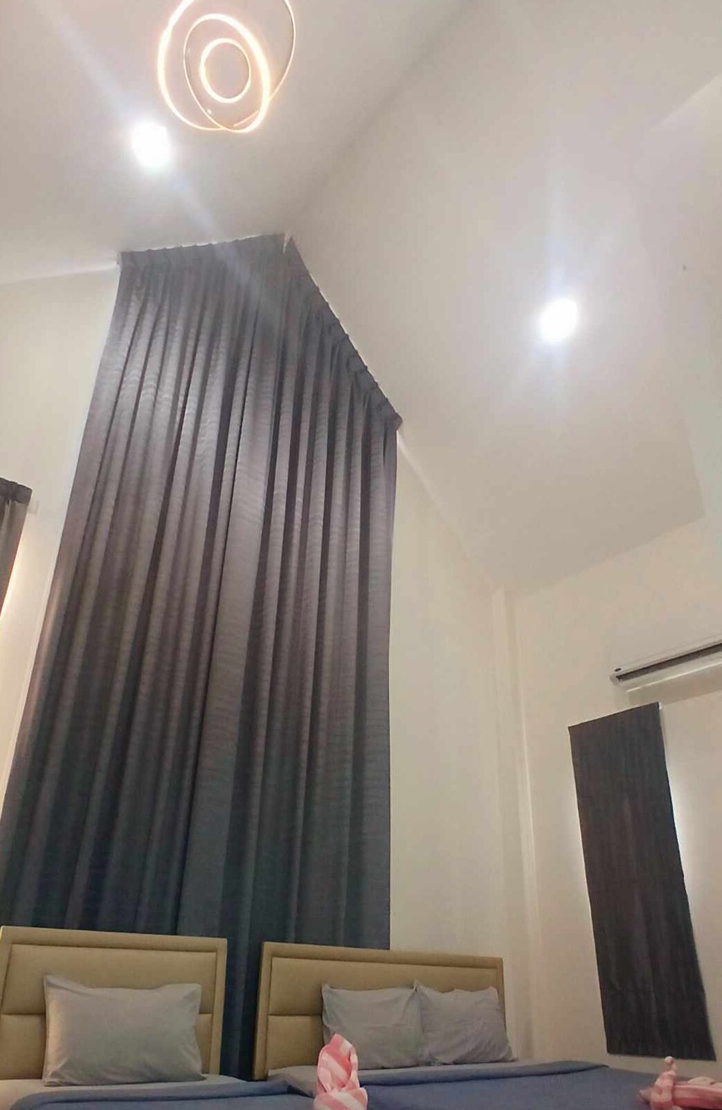 Quarto de hotel com uma cama grande, duas almofadas e cabeceira. Há uma cortina na janela e um lustre no teto.