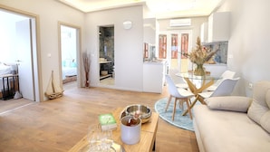 A modern living room with a wooden floor, a glass coffee table, a beige sofa, and a dining area with white chairs and a glass table. - Vostalo apartments by Kassiopi's Sea (Kassiopi)