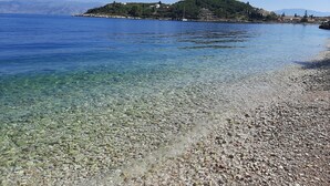 A pebble beach with clear water leading to a hill with buildings. - Vostalo apartments by Kassiopi's Sea (Kassiopi)