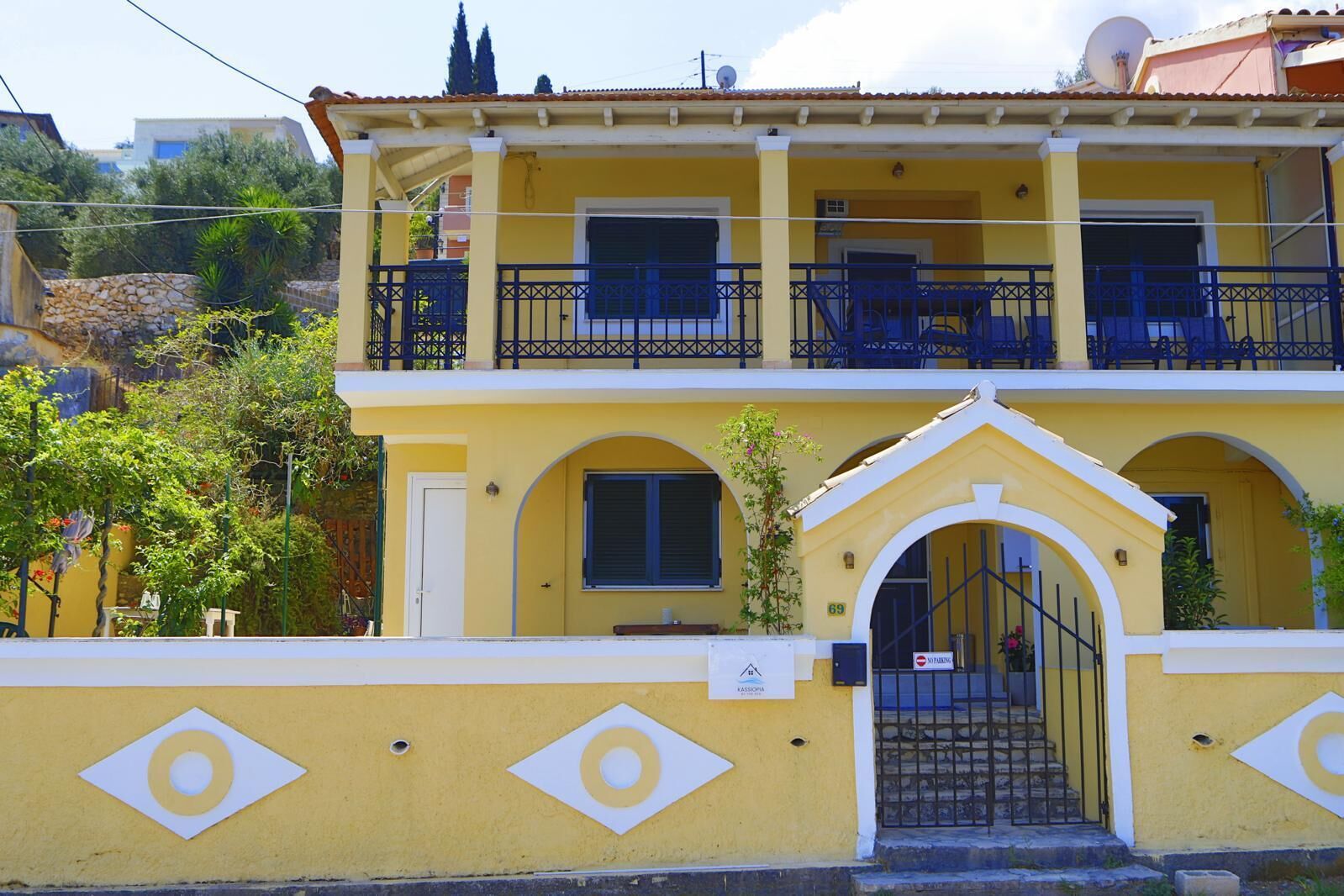 A two-story yellow building with a gated entrance, balconies, and a small garden area.