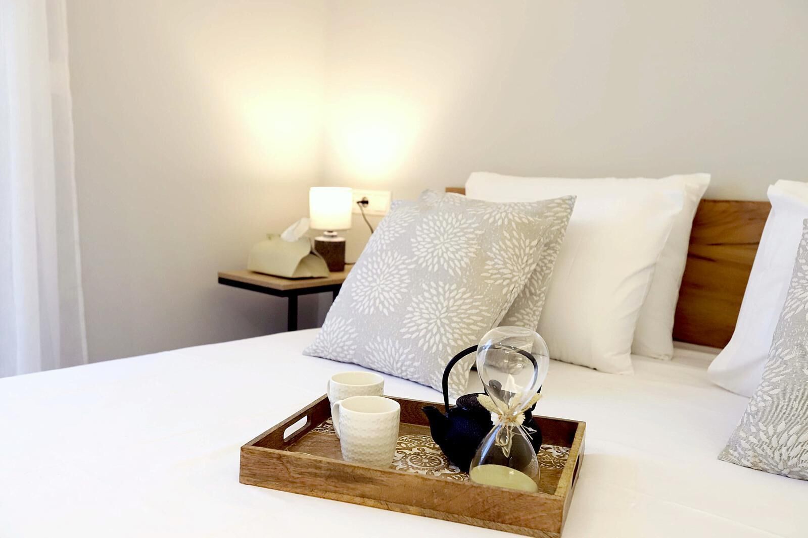 A neatly made bed with a tray of tea set, a glass, and a candle on a wooden tray.
