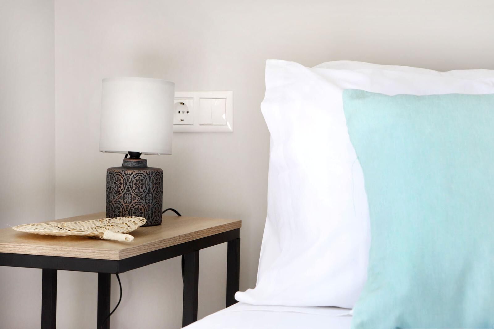 Ein ordentlich eingerichtetes Hotelzimmer mit Nachttisch, Lampe, Steckdose und einem Bett mit weißem und türkisblauem Kissen.