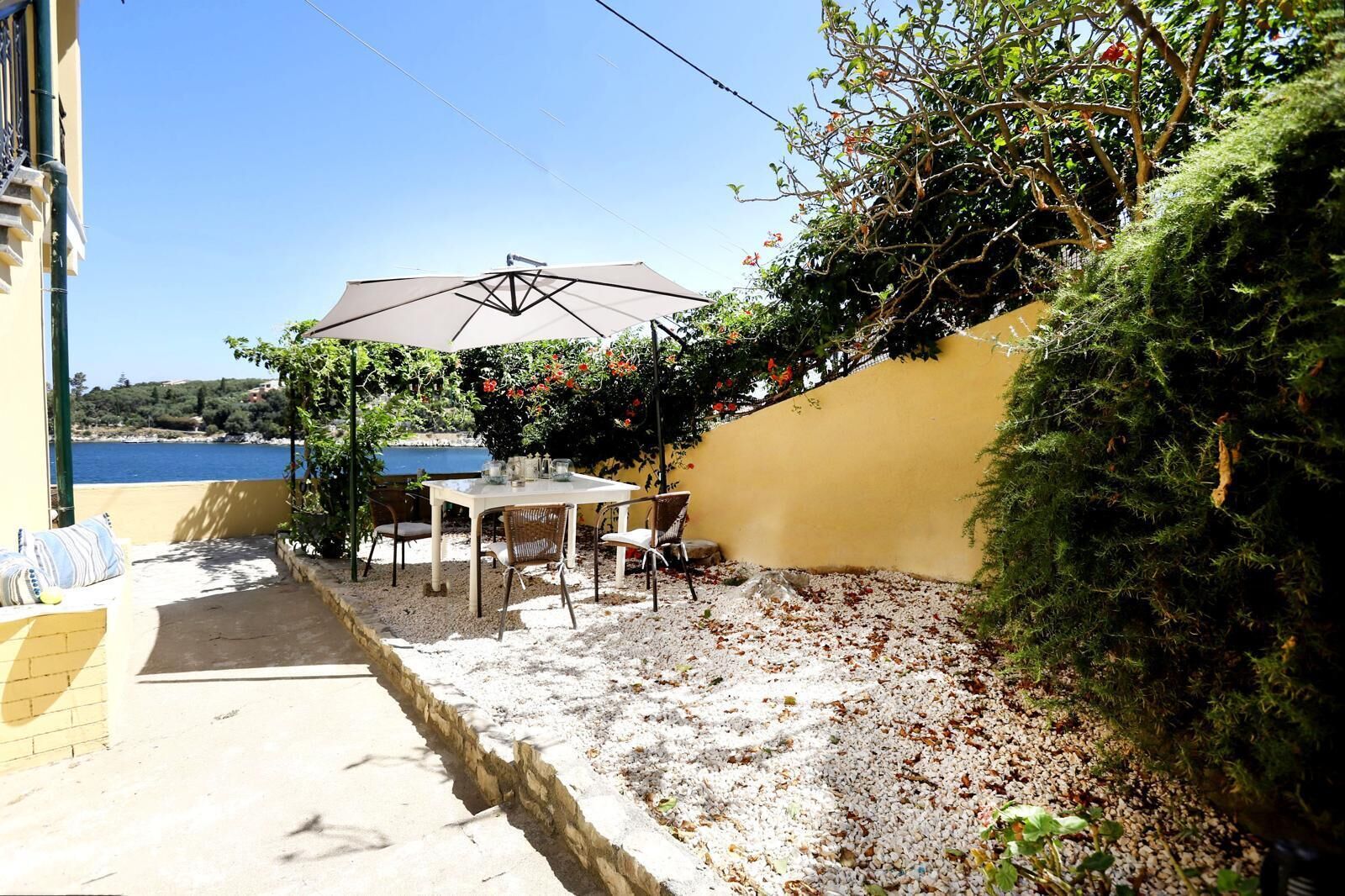 Eine Terrasse mit Tisch und Stühlen, einem weißen Sonnenschirm und Blick auf das Meer.