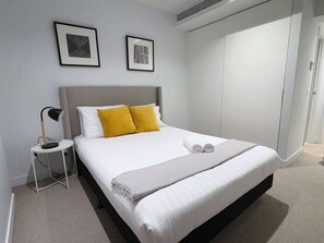 A neatly made bed with white and yellow pillows, a bedside lamp, and a framed picture on the wall. - Bright & Modern 2BR Apt on Iconic Chapel Street (South Yarra)