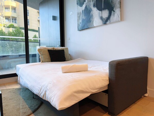 A modern bedroom with a bed, a window with a view of buildings, and a painting on the wall.