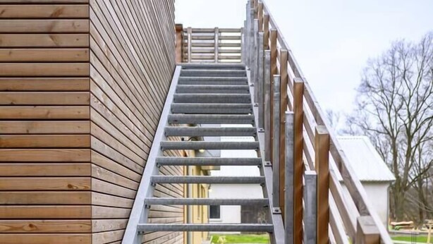 Een metalen trap met houten leuningen die leidt naar een gebouw met horizontale houten bekleding.