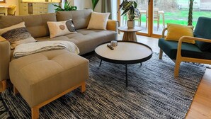 A living room with a sofa, ottoman, coffee table, and chairs. There is a kitchen in the background and a view of the outdoors through large windows. - Pausenhof Spreewald - pond view with terrace (Unterspreewald)