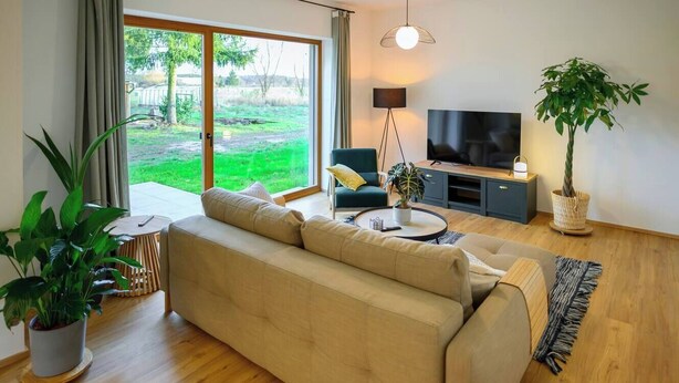 A modern living room with a beige sofa, a television, and a large window overlooking a grassy area. - Pausenhof Spreewald - pond view with terrace (Unterspreewald)