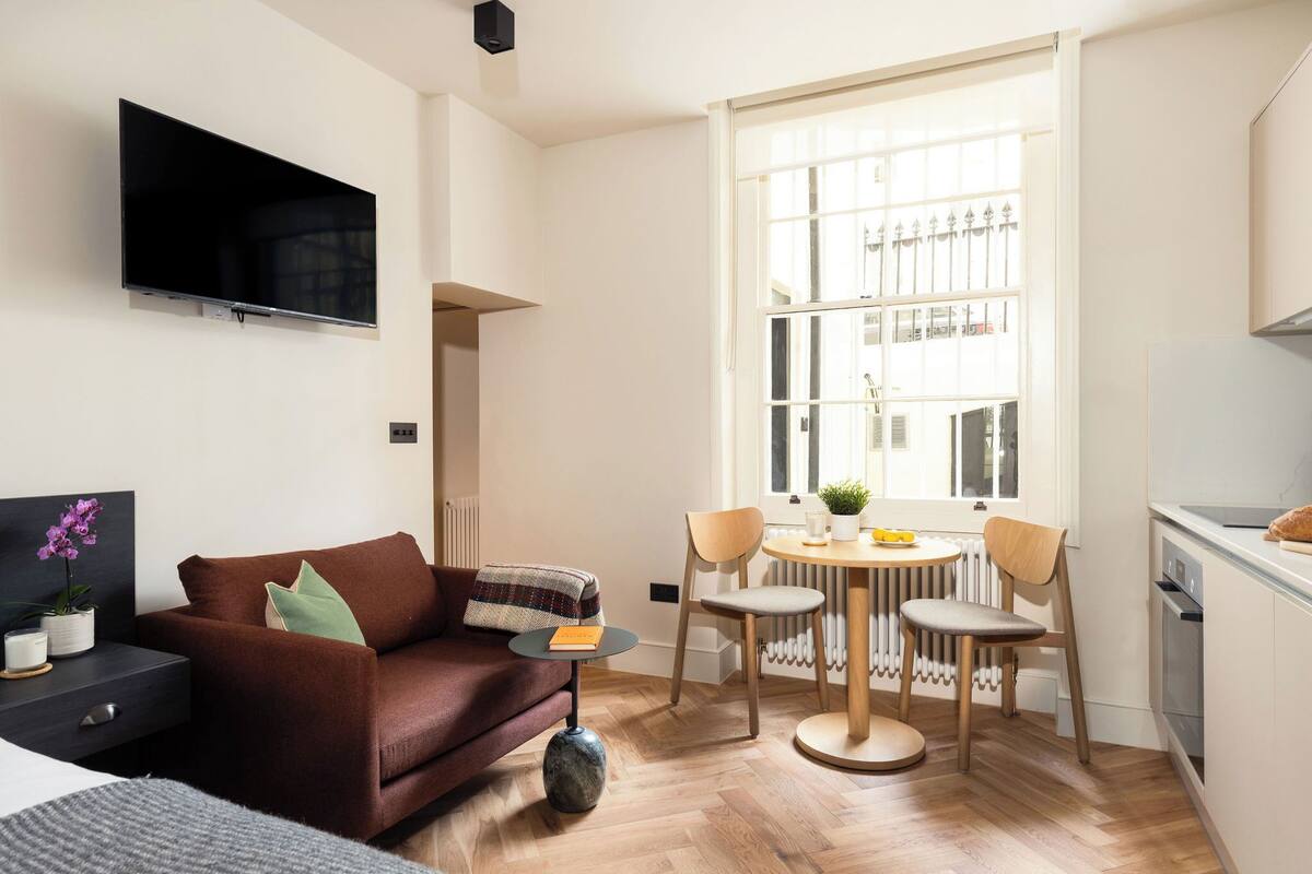 A compact living space with a sofa, a small round table, two chairs, a TV mounted on the wall, and a window with a view of buildings.