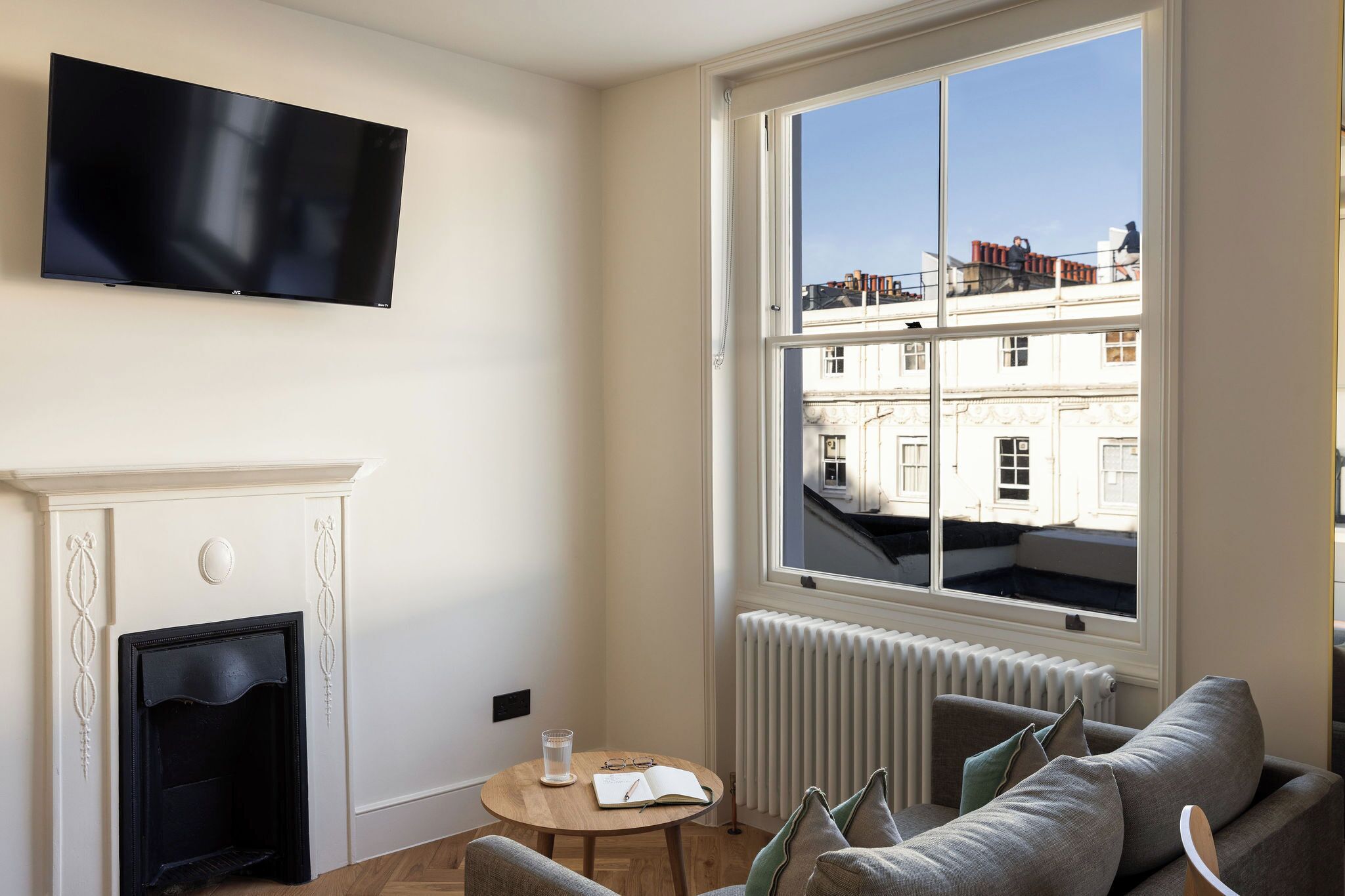 Un salon moderne avec une cheminée, un téléviseur à écran plat fixé au mur, une table ronde en bois sur laquelle sont posés un livre et des lunettes, et une grande fenêtre donnant sur les bâtiments environnants.