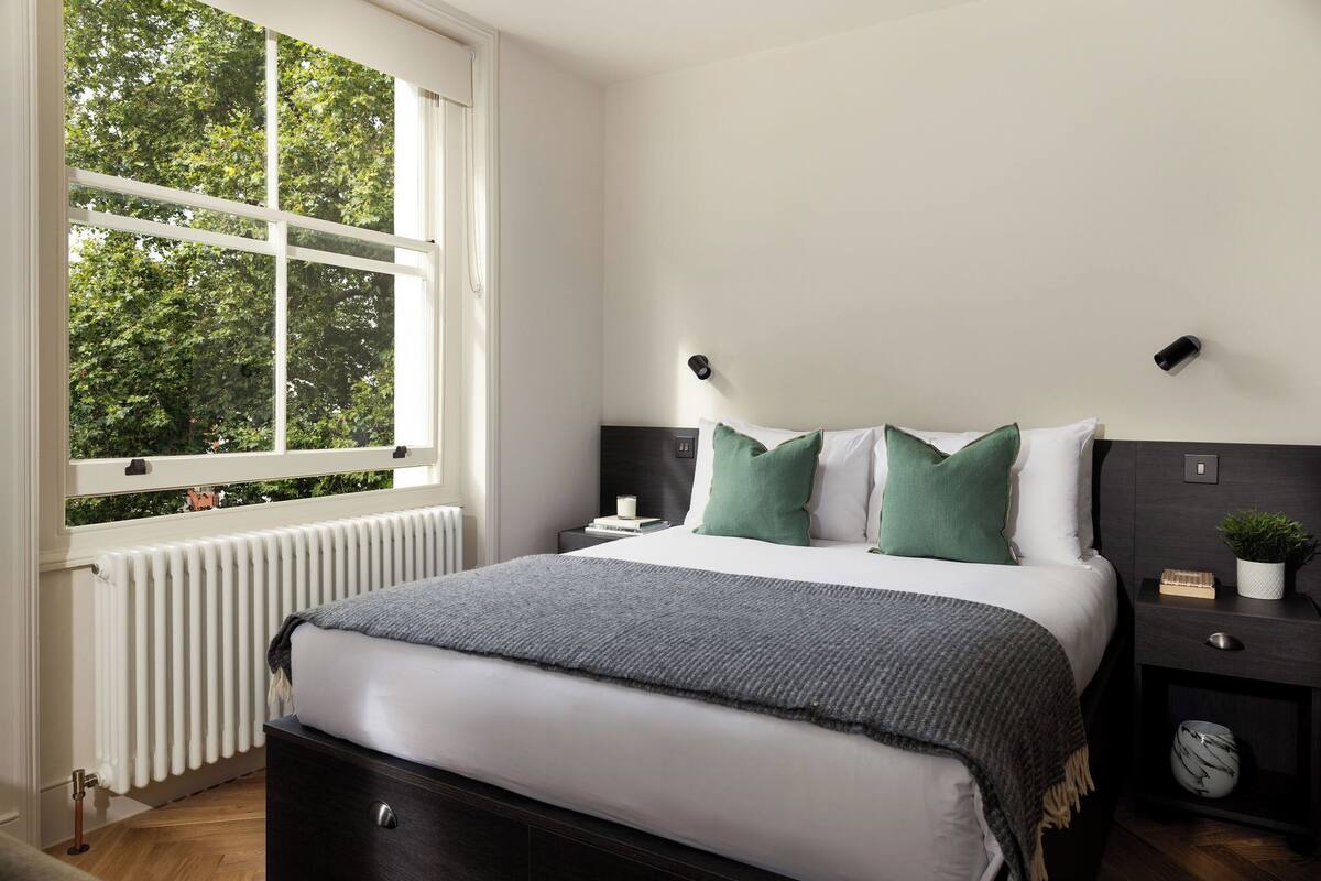A bedroom with a large window, a bed with green pillows, a bedside table with a plant, and a radiator.