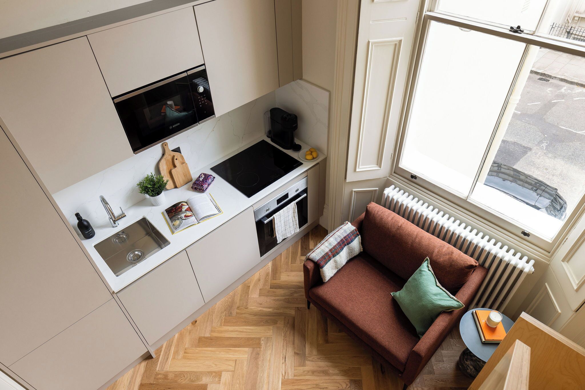 Cucina compatta con forno a microonde, macchina del caffè e lavello. Si vede un divano marrone e un tavolino con un libro e una tazza.