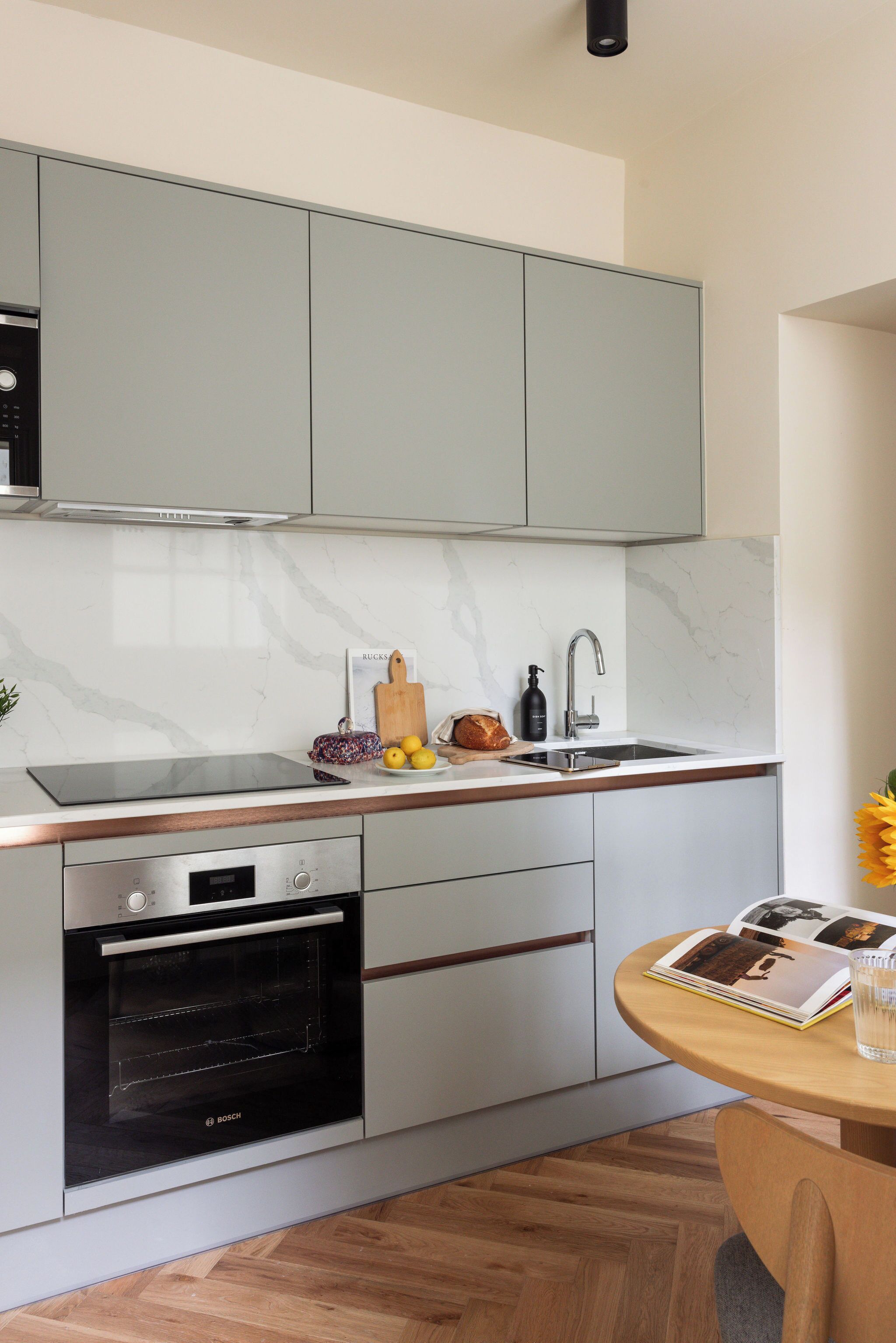 A modern kitchen with grey cabinets, a built-in oven, a sink, and a round wooden table with a magazine and a glass.