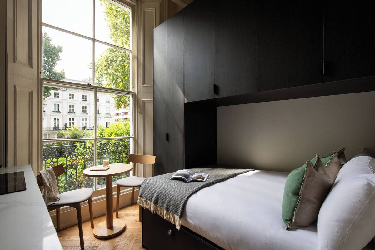 A bedroom with a large window, a bed with pillows, a bedside table, and a chair.